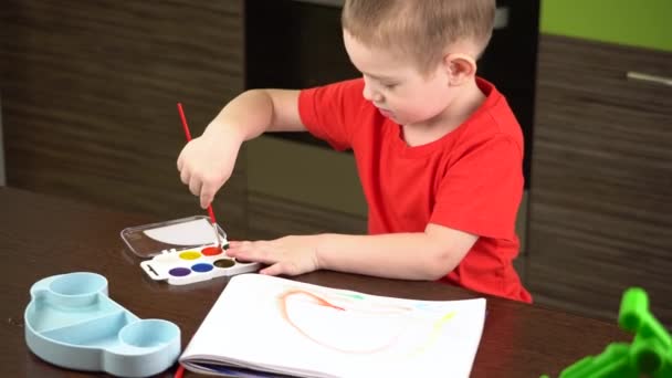 Niño en una camiseta roja pinta con acuarelas de colores en la mesa — Vídeos de Stock