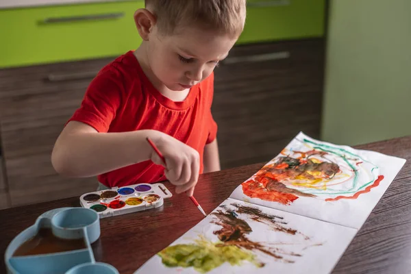 A child in a red T-shirt paints with colorful watercolors at the table