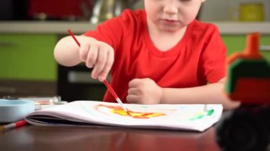 Kırmızı tişörtlü çocuk masada renkli suluboya resimler yapıyor.