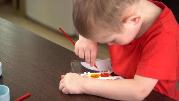 Barn i en röd T-shirt färger med färgglada akvareller vid bordet — Stockvideo