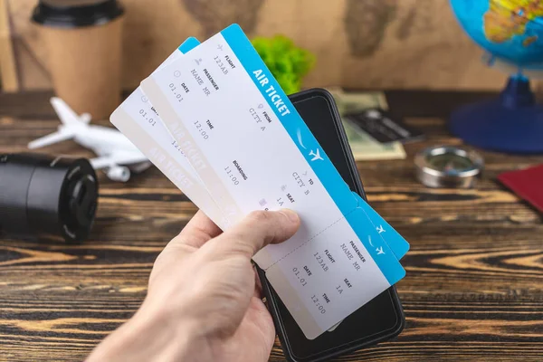 Hand is holding airline tickets and a phone over a wooden table with other travel accessories. Concept of holidays and easily book tickets.