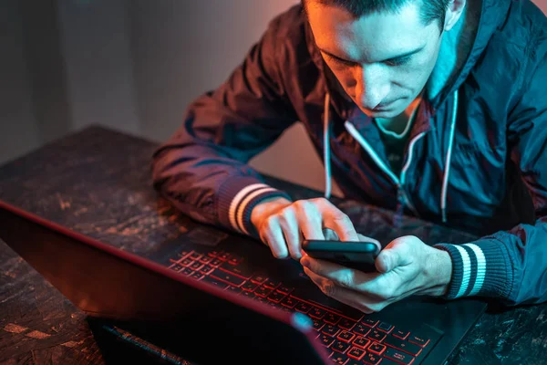 A hacker with a phone is typing on a laptop keyboard in a dark room under a neon light. The concept of cybercrime fraud and identity theft