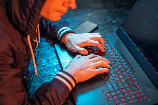 A hooded hacker is typing on a laptop keyboard in a dark room under a neon light. The concept of cybercrime fraud and identity theft