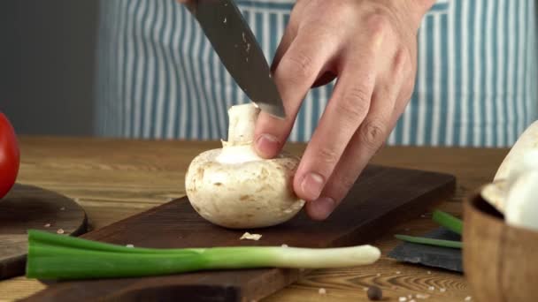 Мужчина-повар в синем фартуке режет свежие грибы ножом. концепция процесса приготовления вкусного блюда — стоковое видео
