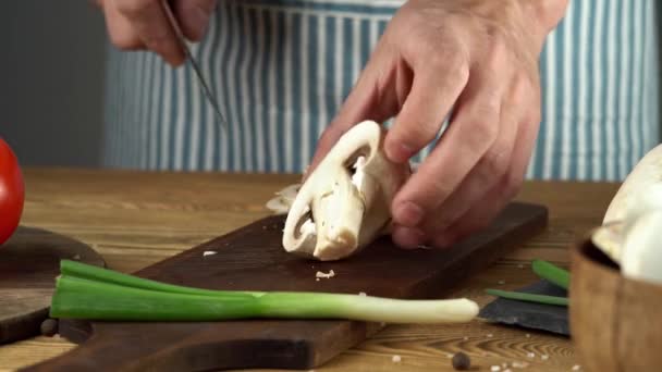Um chef de avental azul corta cogumelos frescos com uma faca. Conceito de processo de cozinhar um prato delicioso — Vídeo de Stock