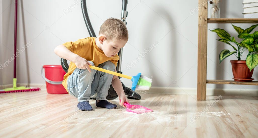 A cute little boy is sweeping the trash from the floor with a broom to a dustpan. Child helps the parents in cleaning the house