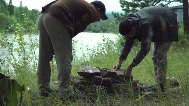 Due escursionisti maschi nella foresta vicino al fiume stanno impilando pietre in una pila per fare un falò per cucinare. — Video Stock