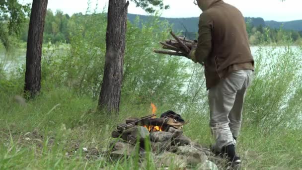 Nehir kıyısındaki ormandaki adam bir kucak dolusu kuru odun alıp ateşe atıyor. — Stok video