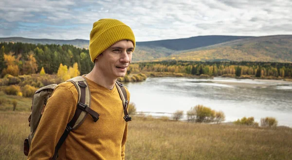 バックパック付きの黄色の帽子とセーターの男の旅行者は 黄金の森と丘に沿って歩いています ハイキング 秋の気分の概念 — ストック写真
