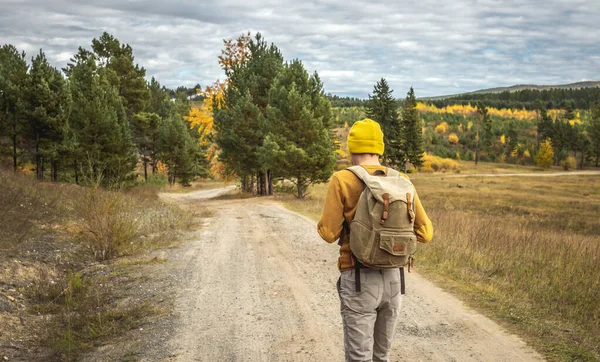 Traveler in a yellow hat and a sweater with a backpack is walking on an empty road along autumn golden forest. Concept of freedom, travel, hiking and autumn mood