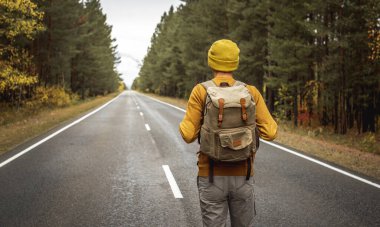 Sarı şapkalı ve sırt çantalı bir kazaklı gezgin sonbahar ormanı boyunca boş düz bir yolda yürüyor. Özgürlük kavramı, seyahat, yürüyüş ve sonbahar havası