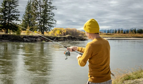 黄色のセーターを着た若い漁師と帽子は 彼の手に回転棒で秋の森の川岸で釣りをしています リラックスした屋外レクリエーション ライフスタイル 趣味の概念 — ストック写真