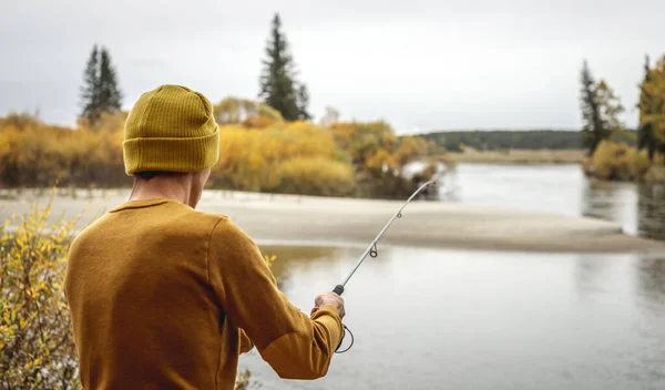 黄色のセーターを着た若い漁師と帽子は 彼の手に回転棒で秋の森の川岸で釣りをしています リラックスした屋外レクリエーション ライフスタイル 趣味の概念 — ストック写真