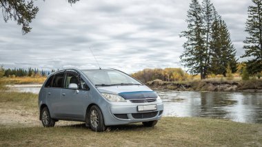Araba hızlı bir nehrin kıyısında, sarı yeşil bir sonbahar ormanının yanında park halinde. Seyahat kavramı, güzel doğa ve sonbahar havası.