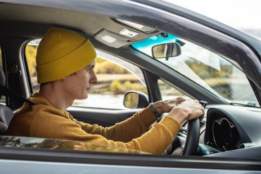 Sarı şapkalı ve kazaklı bir adam nehir boyunca ve güzel sonbahar ormanlarında araba sürüyor. Doğa kavramı, seyahat ve sonbahar havası