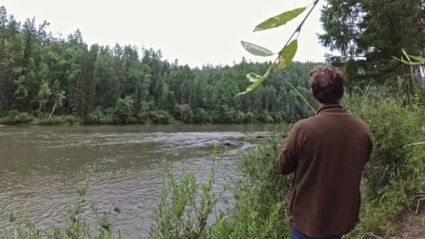 Homem está de pé na margem do rio com uma vara de pesca em suas mãos e captura de peixe — Vídeo de Stock