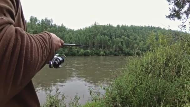 Człowiek stoi na brzegu rzeki z wędką w rękach i łapie ryby. — Wideo stockowe
