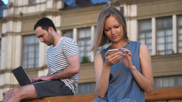 Mężczyzna za pomocą laptopa i kobieta za pomocą smartfona siedzi na ławce o — Zdjęcie stockowe