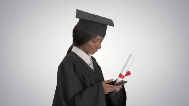 Mosolygó afro-amerikai női diplomás diploma sms-ezés a telefonján, miközben séta gradiens háttér. — Stock videók