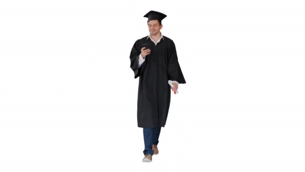 Estudiante masculino en vestido de graduación revisando su teléfono mientras camina sobre fondo blanco. — Vídeos de Stock