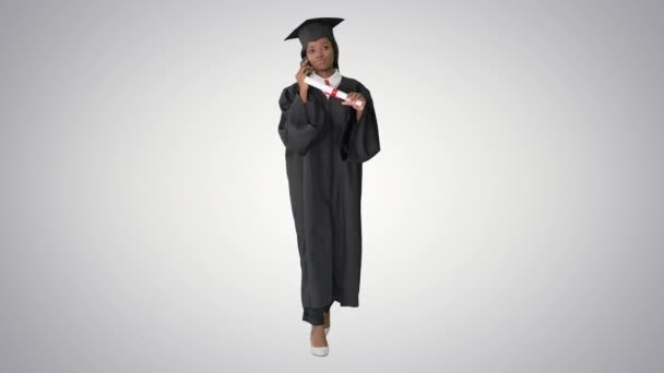 Αφρο-Αμερικανίδα φοιτήτρια με στολή αποφοίτησης να μιλάει στο τηλέφωνο ενώ περπατούσε σε επίπεδο φόντου. — Αρχείο Βίντεο