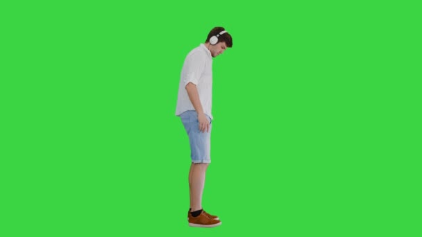在一个绿色的屏幕上，Chroma键上散步时，一个随随便便的年轻人一边用耳机听音乐. — 图库视频影像