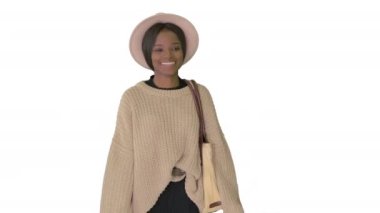 Beyaz arka planda yürürken örgü ve beyaz şapkalı gülümseyen Afro-Amerikan kadın..