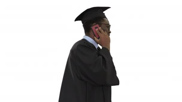 Sonriente afroamericano estudiante masculino en bata de graduación hablando por teléfono mientras camina sobre fondo blanco. — Vídeos de Stock