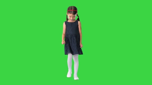 Nettes kleines Mädchen mit Zöpfen in schwarzem Kleid, das zu ihren Füßen hinunterschaut und dann auf einem Green Screen in die Kamera lächelt, Chroma Key. — Stockvideo