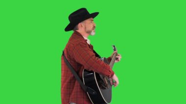 Kovboy şapkalı bir adam yeşil ekranda yürürken şarkı söylüyor ve gitar çalıyor..