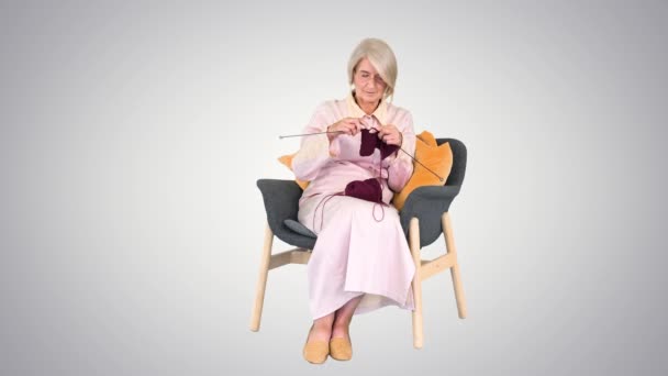 Dama de edad inteligente tejiendo sentado en una silla sobre fondo degradado. — Vídeo de stock