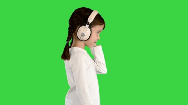 Niña vestida de blanco caminando con auriculares y asintiendo con la cabeza al ritmo de la música en una pantalla verde, Chroma Key. — Vídeos de Stock