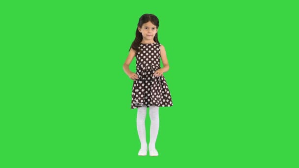 Dunkelhaariges kleines Mädchen im gepunkteten Kleid, das eine schwarze Sonnenbrille aufsetzt und auf einem Green Screen in die Kamera winkt, Chroma Key. — Stockvideo