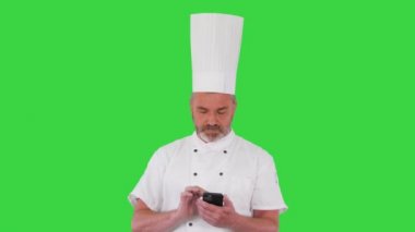 Beyaz üniformalı olgun bir aşçı yeşil ekranda, Chroma Key 'de cep telefonuna mesaj atıyor..