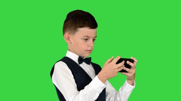 Koncentrerad pojke i vit skjorta, fluga och väst tittar på något på sin telefon på en grön skärm, Chroma Key. — Stockvideo