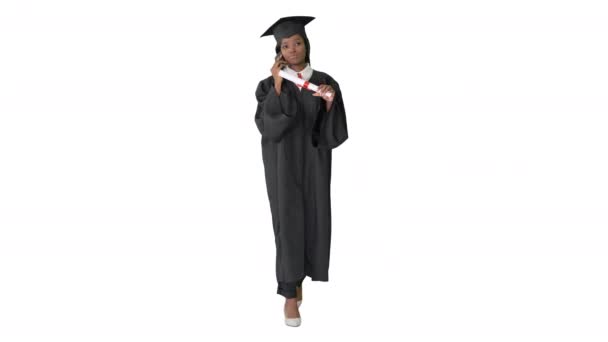 Αφρο-Αμερικανίδα φοιτήτρια με στολή αποφοίτησης να μιλάει στο τηλέφωνο ενώ περπατάει σε λευκό φόντο. — Αρχείο Βίντεο