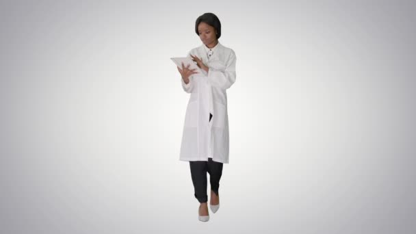 Afro-Américaine médecin tenant un ordinateur tablette tout en marchant sur fond de dégradé. — Video