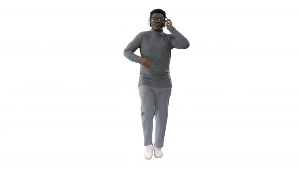 Uśmiechnięty Afroamerykanin trzymający słuchawki jedną ręką i wbijający się w muzykę podczas chodzenia przed kamerą na białym tle. — Wideo stockowe
