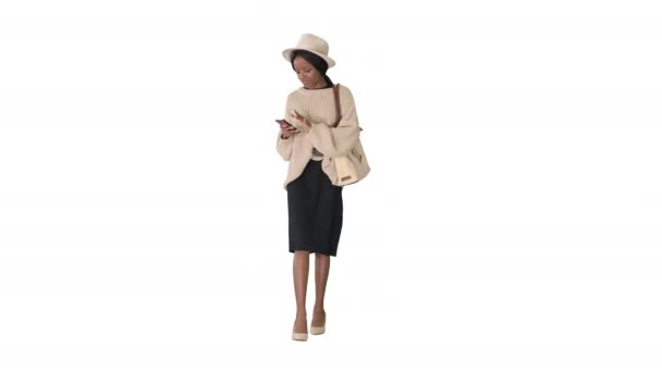 Elegante donna afroamericana in maglieria e cappello utilizzando il telefono mentre cammina su sfondo bianco. — Video Stock