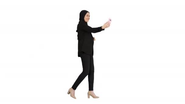 Wanita muslim muda dalam jilbab memiliki panggilan video di teleponnya saat ia berjalan di atas latar belakang putih. — Stok Video
