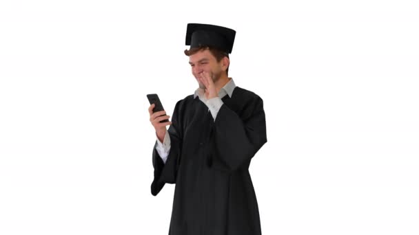 Śmiejący się student w sukni dyplomowej i zaprawy mając wideo połączenie na telefonie na białym tle. — Wideo stockowe