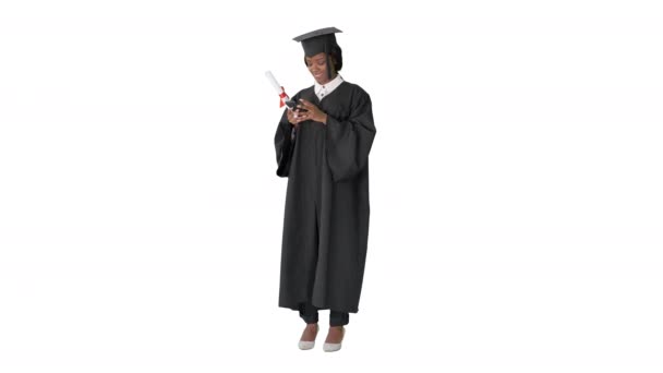 Feliz mujer afroamericana graduada sosteniendo diploma y mensajes de texto en su teléfono sobre fondo blanco. — Vídeos de Stock