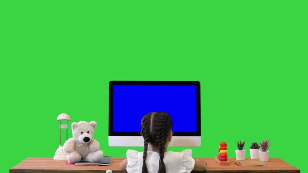 Маленька дівчинка сидить за столом і спостерігає щось на моніторі Mock-up Display on a Green Screen, Chroma Key. — стокове відео