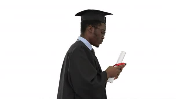 Afričtí američtí muži student v absolventském županu textování na telefonu při chůzi se svým diplomem na bílém pozadí.