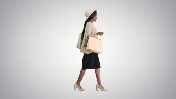 Heureuse femme afro-américaine en bonnet et tricot marchant avec des sacs à provisions sur fond dégradé. — Video