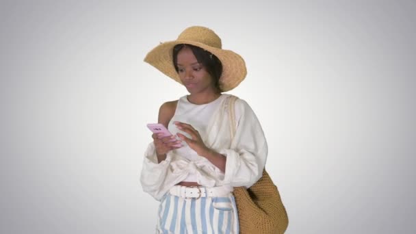 Uśmiechnięta młoda Afroamerykanka w słomkowym kapeluszu smsująca przez telefon na gradientowym tle. — Wideo stockowe