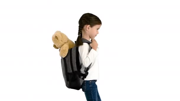 Petite fille mignonne marchant à l'école avec un ours en peluche qui sort de son sac à dos sur fond blanc. — Video