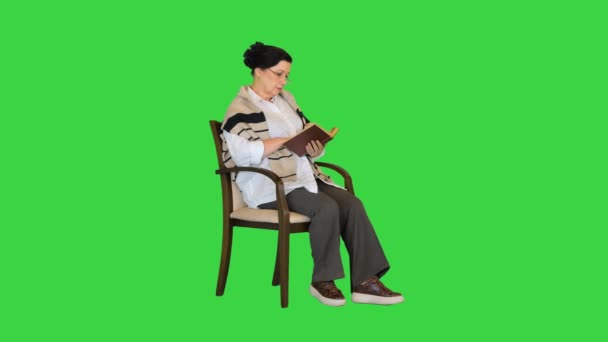 Дружелюбная пожилая женщина, сидящая в кресле и читающая книгу на зеленом экране, Chroma Key. — стоковое видео