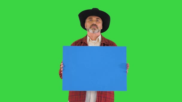 Seriös man i cowboy hatt tittar till kameran och håller tomma plakat på en grön skärm, Chroma Key. — Stockvideo
