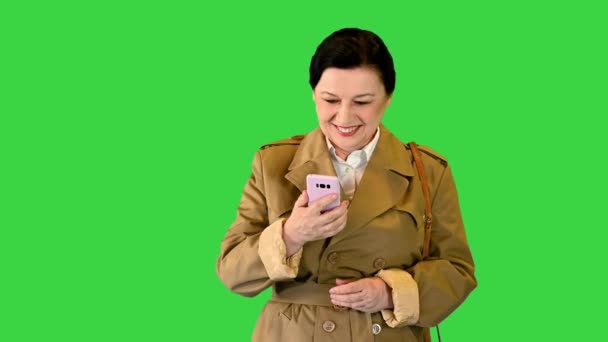 Trençkotlu orta yaşlı kadın yürüyor ve yeşil ekranda akıllı telefon kullanıyor, Chroma Key. — Stok video